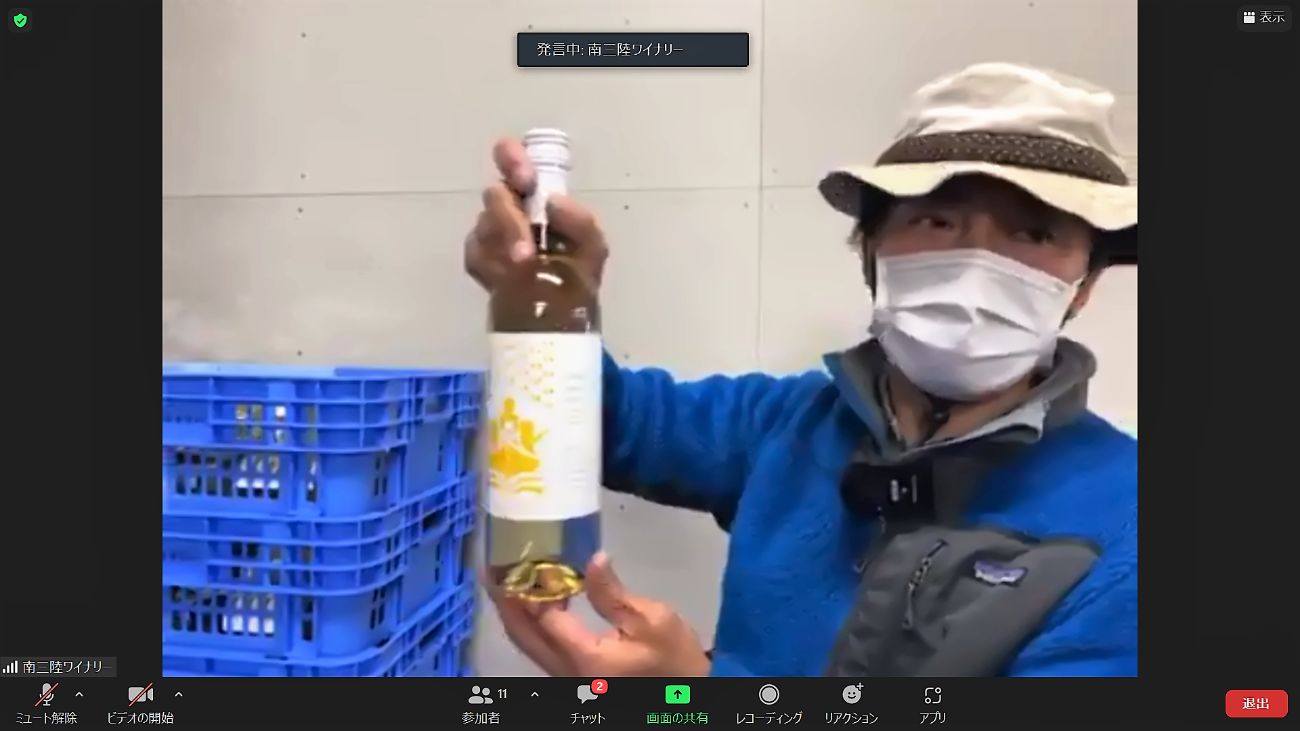 帽子を被り青い服とマスクを着けた男性が、青いケースの中に入っていた白ワインのボトルをカメラに向けて持ち上げて説明しているZoom画面の写真