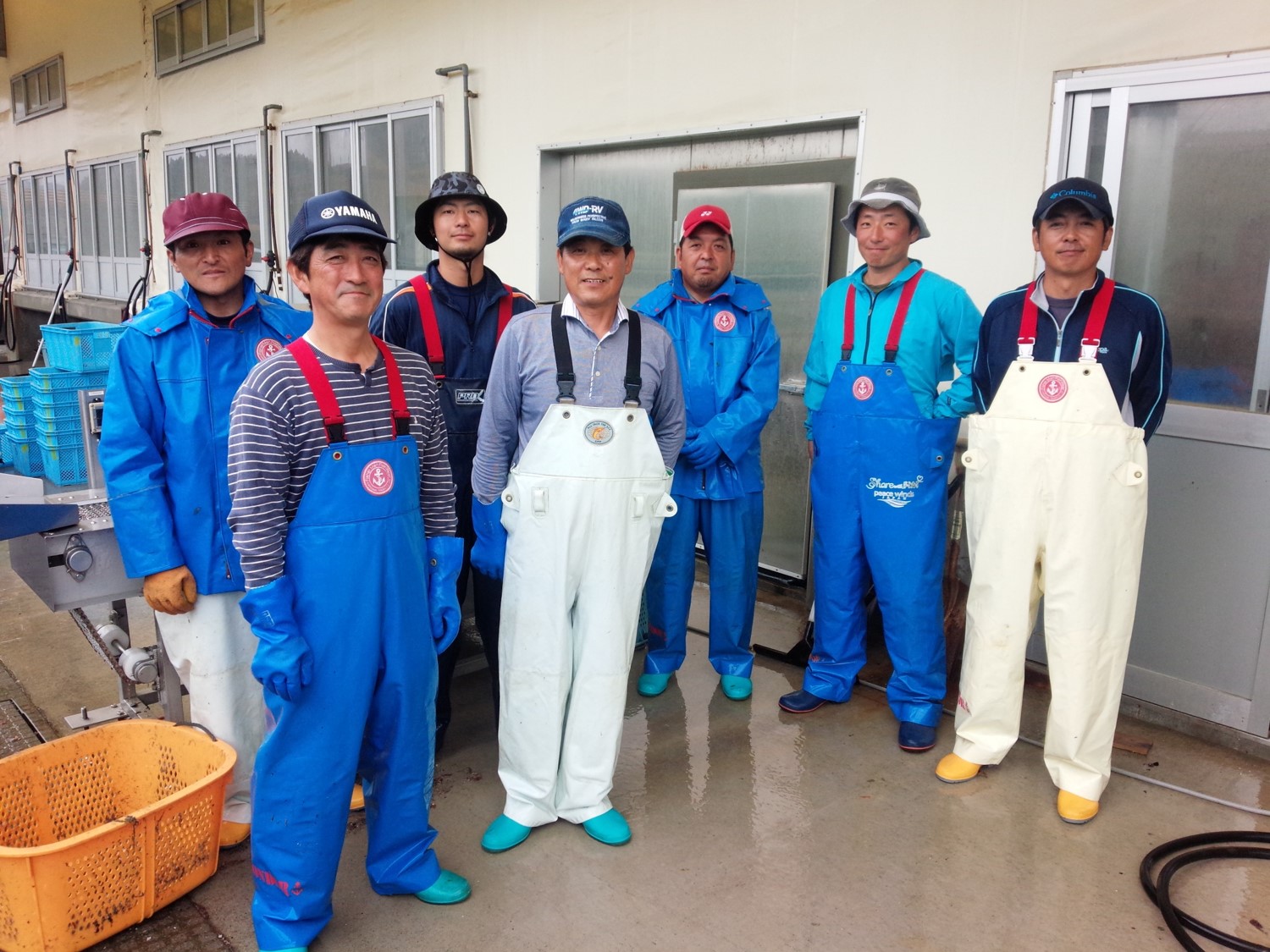 青や白の作業着を着た8人の「あまころ牡蠣」の生産者の男性たちが建物の前に並んで立っている写真