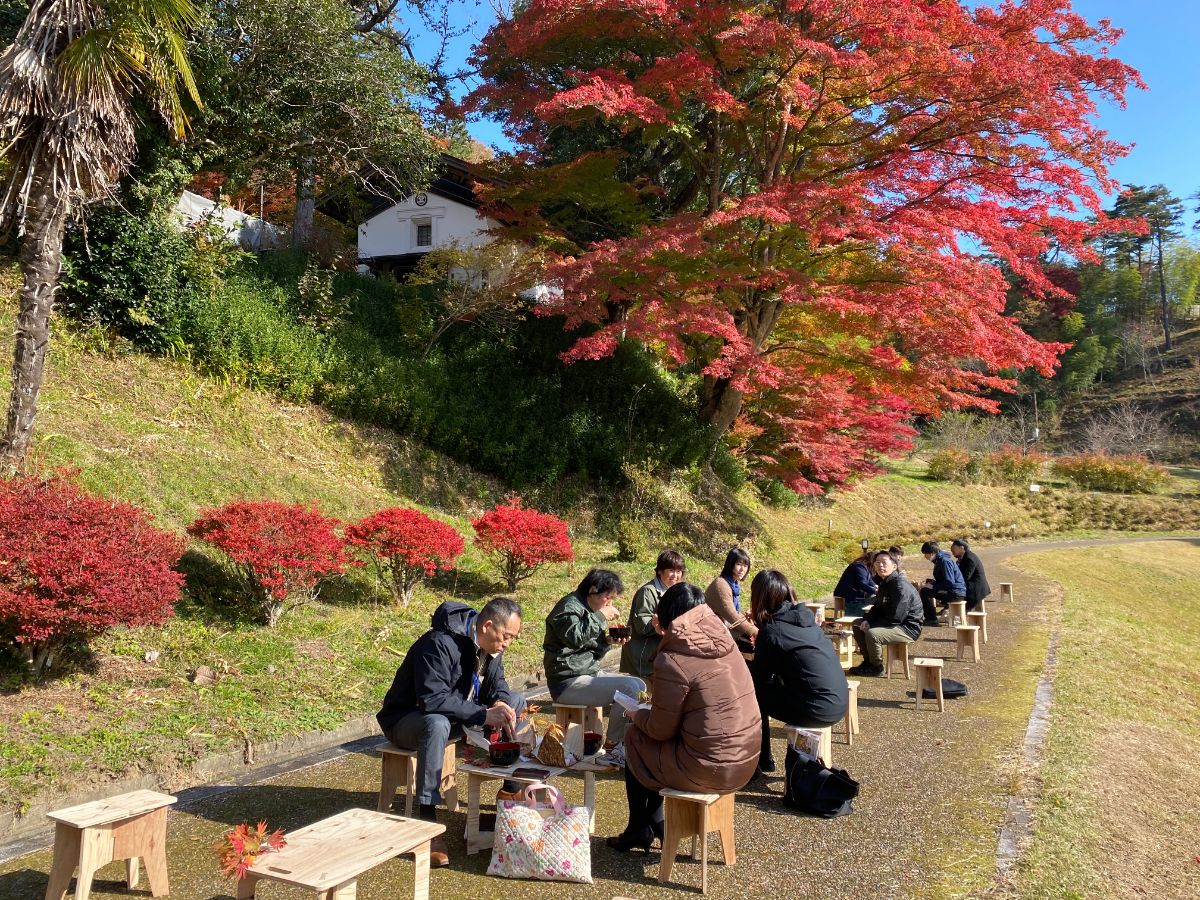 赤く色づいた紅葉の木の側で、木製の椅子とテーブルに座って複数の人々が食事を楽しんでいる写真
