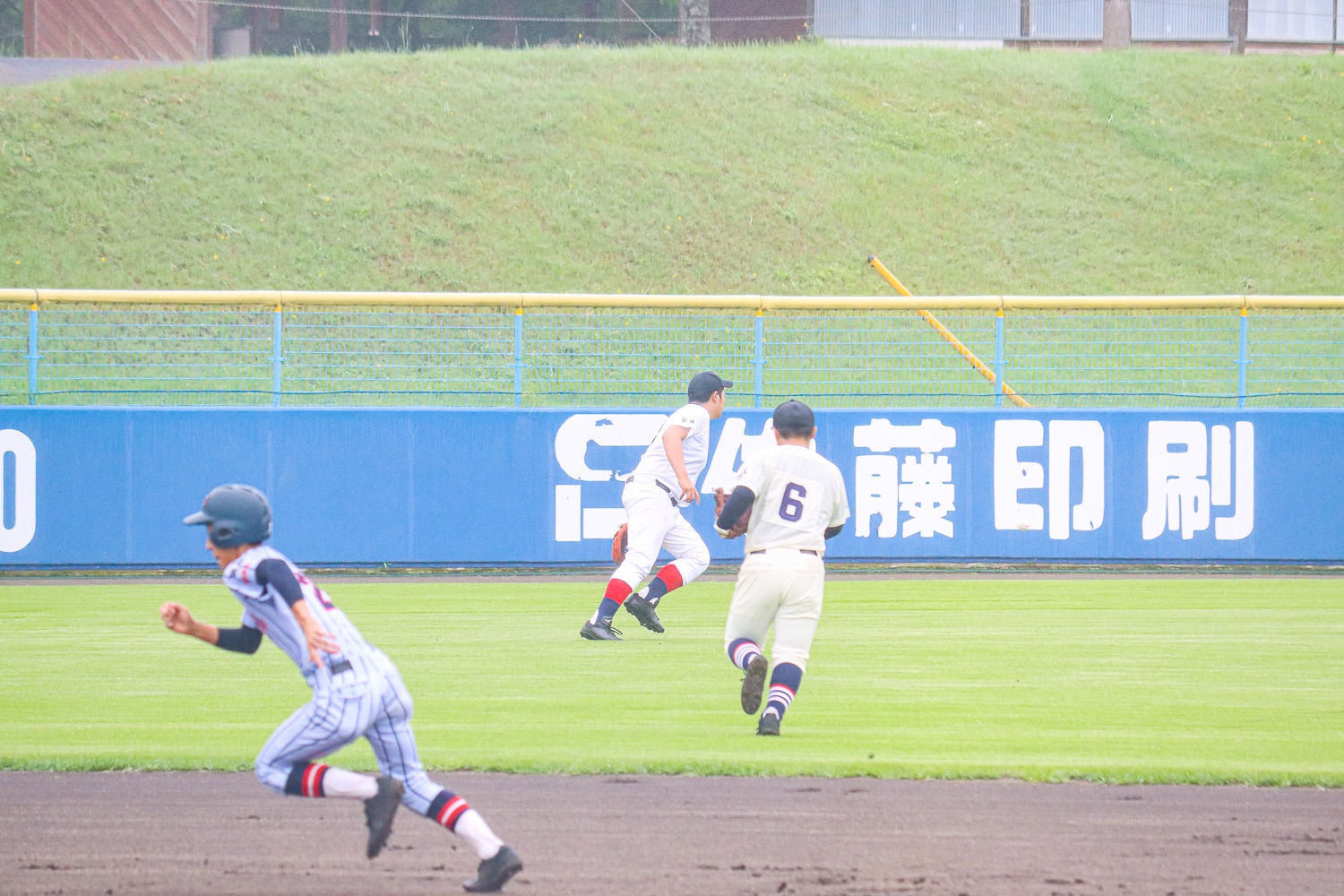 青と白のストライプユニフォームの走者が内野を全力で走る一方、白いユニフォームを着た2人の外野手がフェンス近くで打球を追って走っている様子の写真