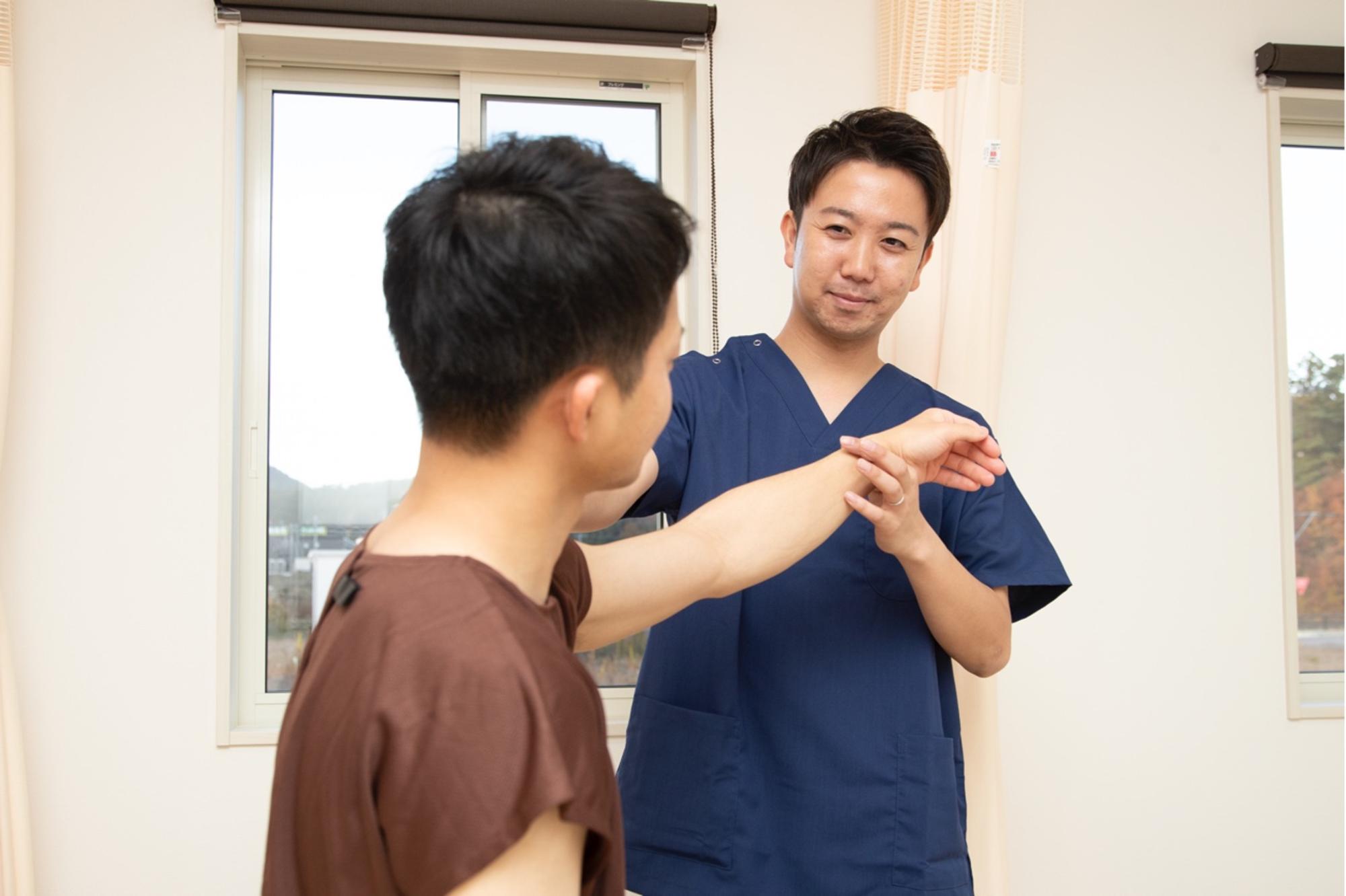 施術着を着た長田さんが室内で患者の腕を丁寧に持ち上げながら状態を確認しており、明るい治療室の中で真剣に施術を行う様子の写真