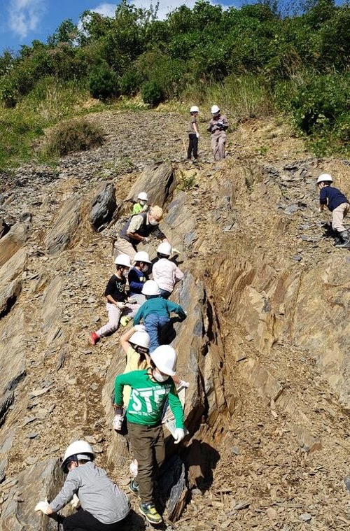 子どもたちが白いヘルメットをかぶって岩山を登りながら地層を観察している様子を写した自然体験学習の写真