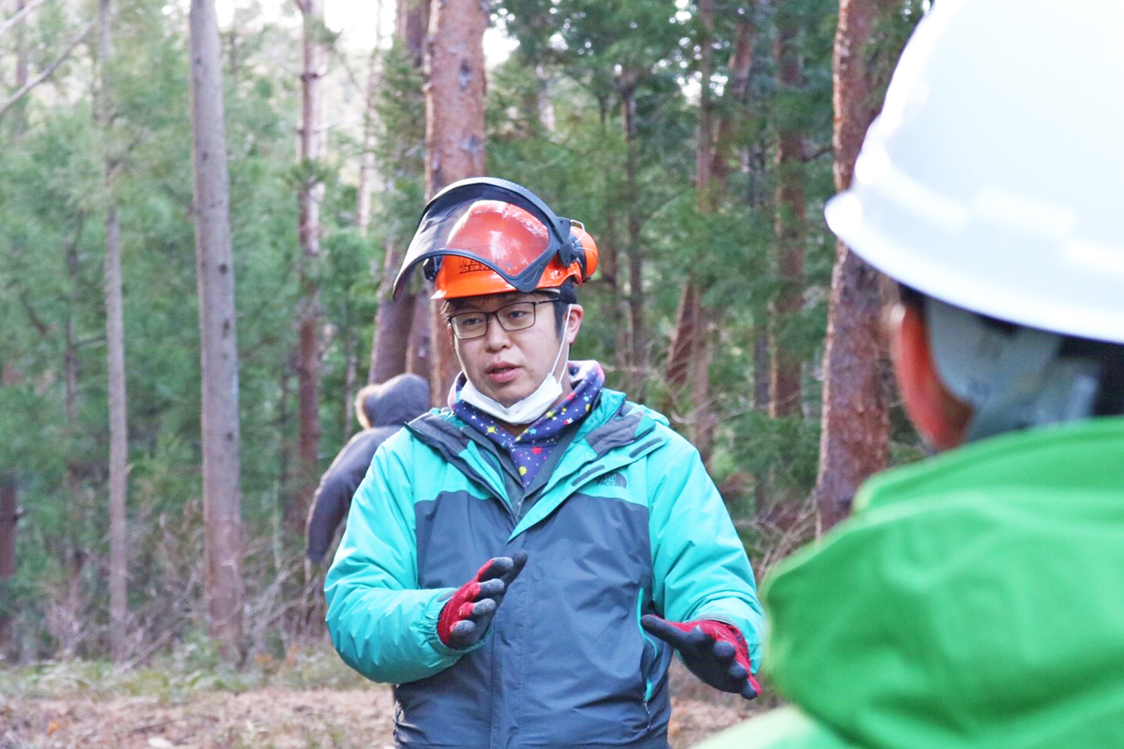 森林の中で防護ヘルメットをかぶった佐藤さんが向かい合って立った人物に話をしている様子の写真