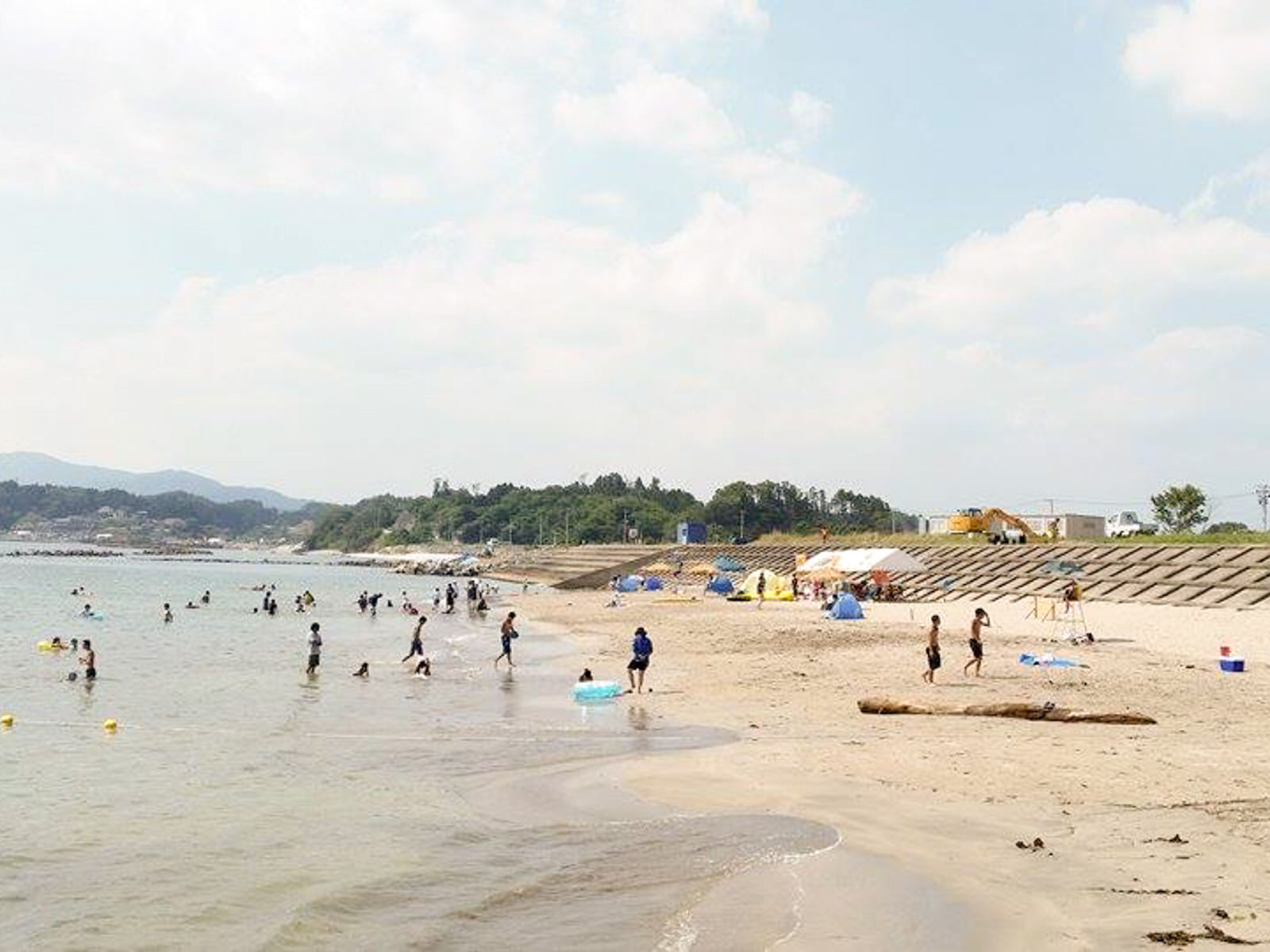 テントや建物、山々が見える明るい昼間の風景が広がっている海水浴場で、たくさんの人が海に入ったり砂浜で遊んだりしている長須賀海水浴場の写真