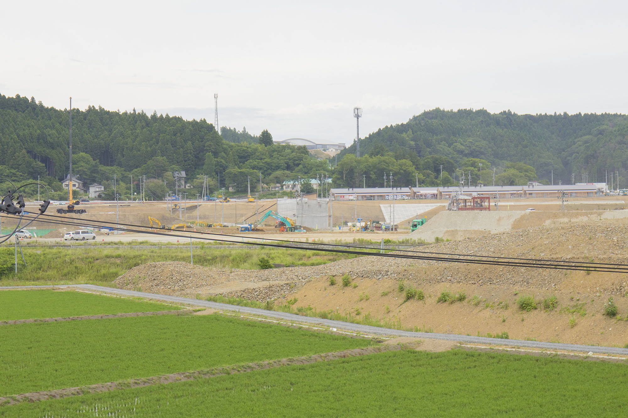 手前に田んぼが広がり、その奥では造成工事が進められており、さらに遠くには森林と山が見える風景を撮影した写真