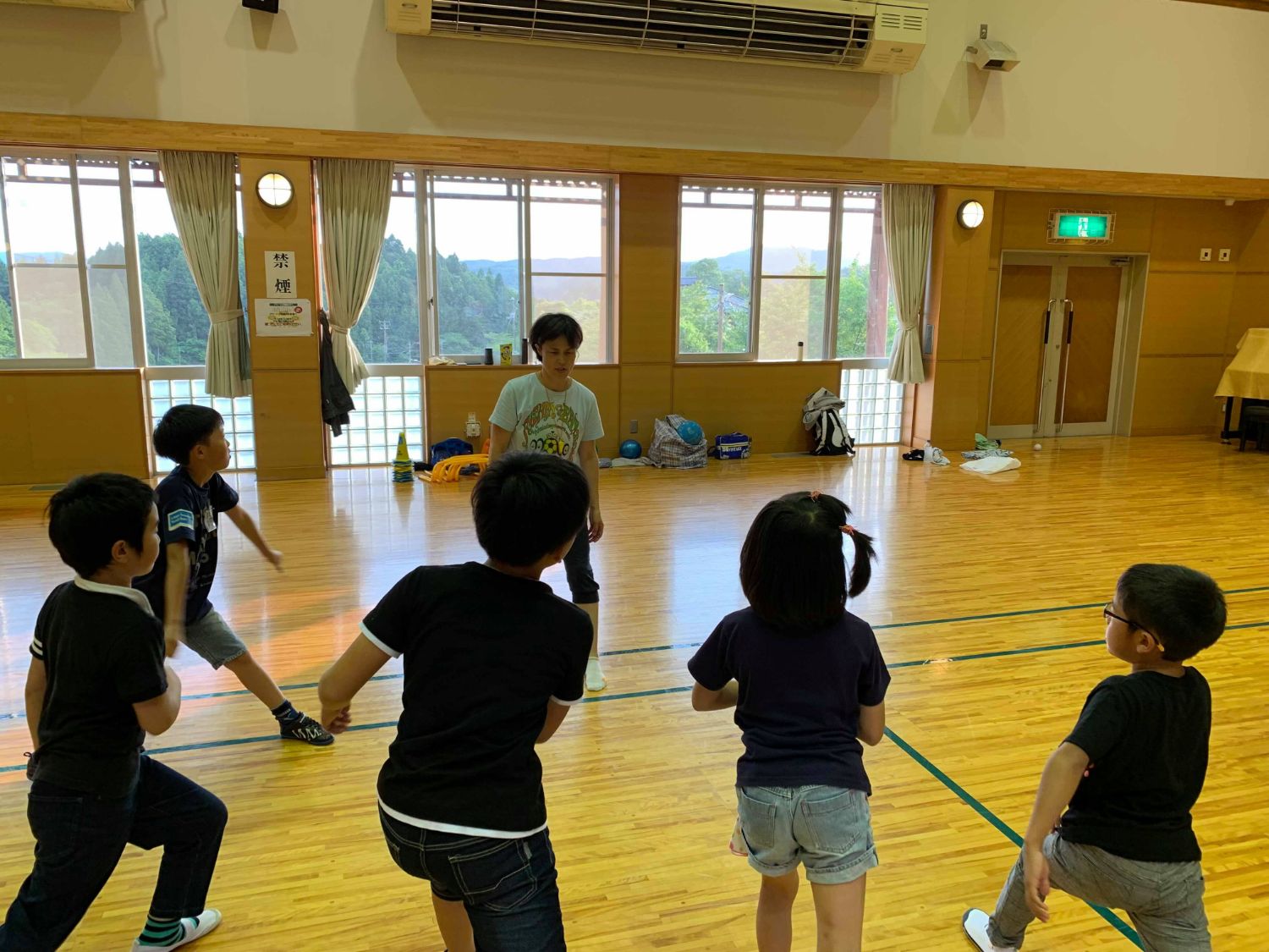 体育館の真ん中で、Tシャツを着た女性の前に半円形に集まり、片足を揚げたり、足を前後に開いたりして運動の指導を受けている子どもたちの写真