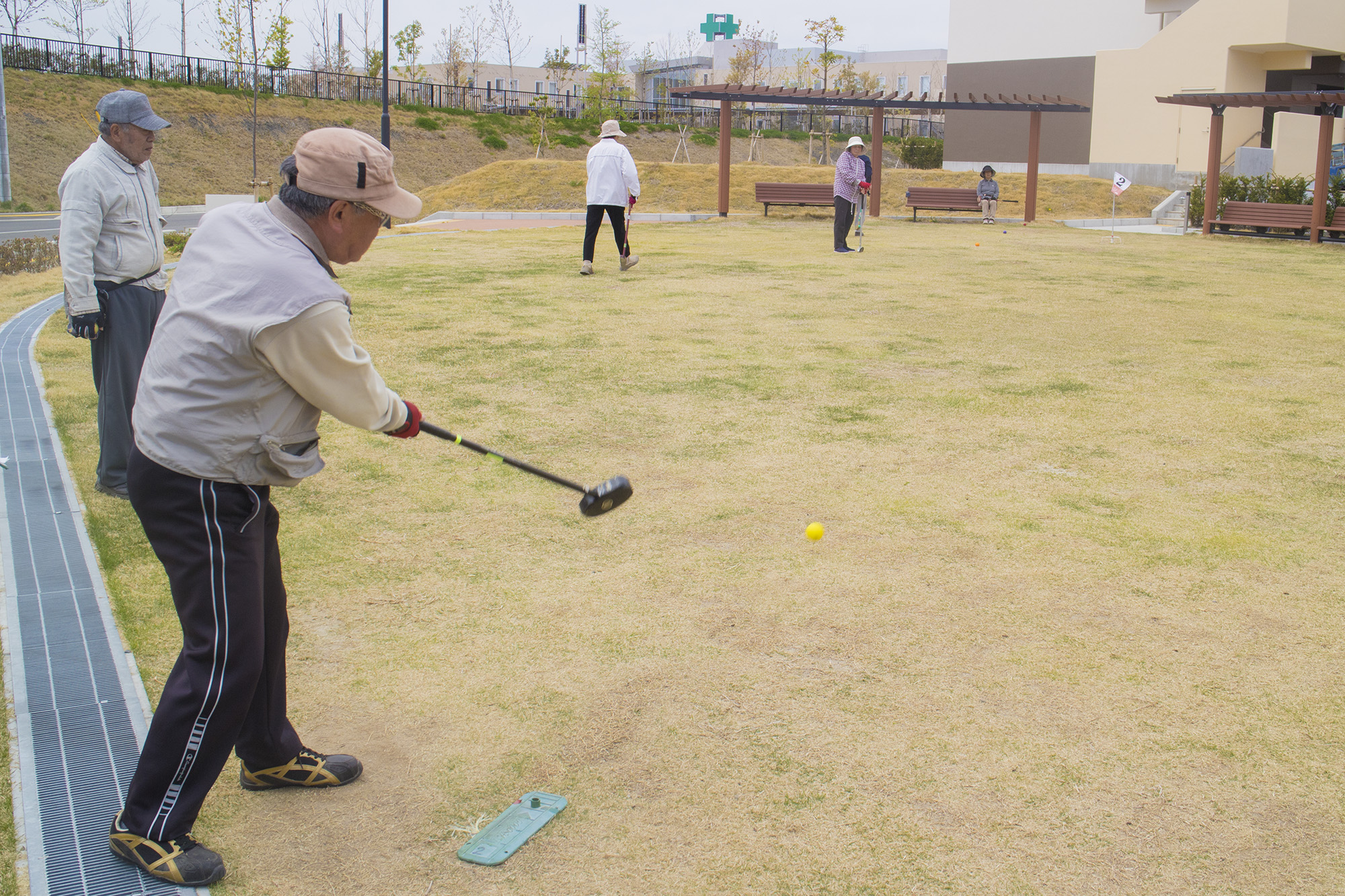 芝生の広場で数人の高齢者がグラウンドゴルフを楽しんでおり、手前ではベージュの帽子をかぶった男性が黄色いボール打った瞬間が捉えられている写真