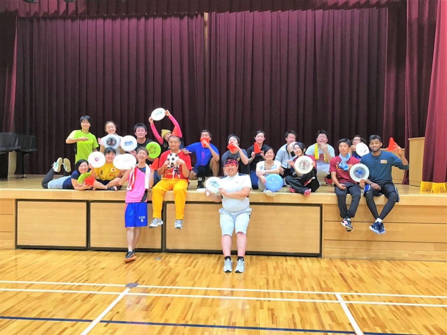 背景にはえんじ色のカーテンがあり、床は白線が引かれ磨かれた木製である体育館のステージ上で、複数の人物が座ったり立ったりしながら、それぞれ様々な色のフリスビーを手に持ってポーズを取っている様子を写している集合写真