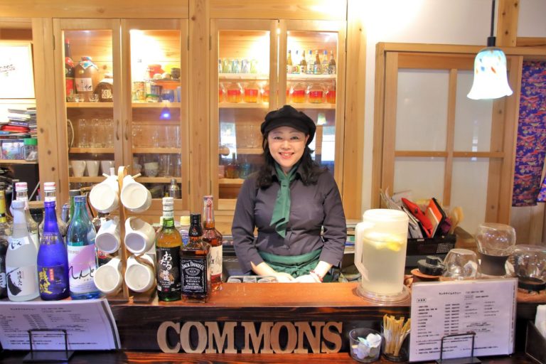 木製の棚に様々な飲み物やグラスが並び、カウンターにはボトルやメニュー表が置かれている飲食店の店内でカウンターの前に黒い帽子をかぶり、紺色の服と緑のスカーフを着けた内海さんが笑顔でたっている写真