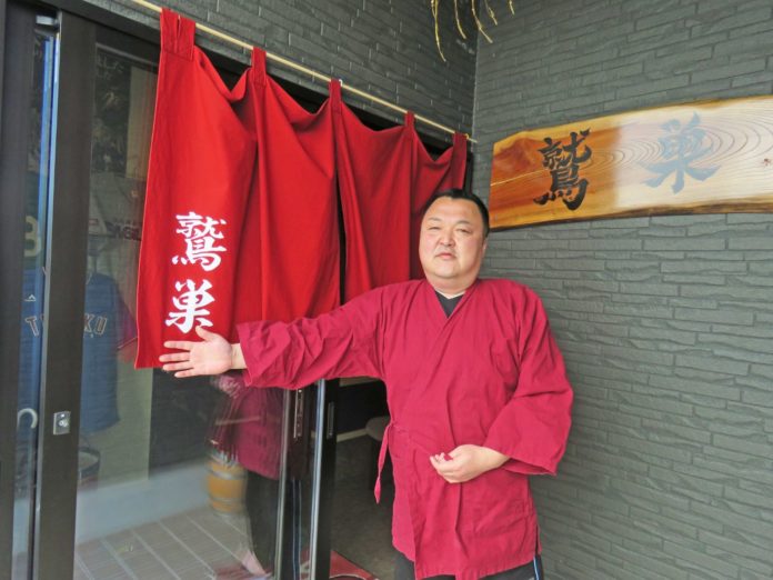 「鷲巣」と書かれた看板や赤い暖簾が設置された居酒屋の入口の前に、赤い作務衣を着た店主の男性が立っている様子を写している写真