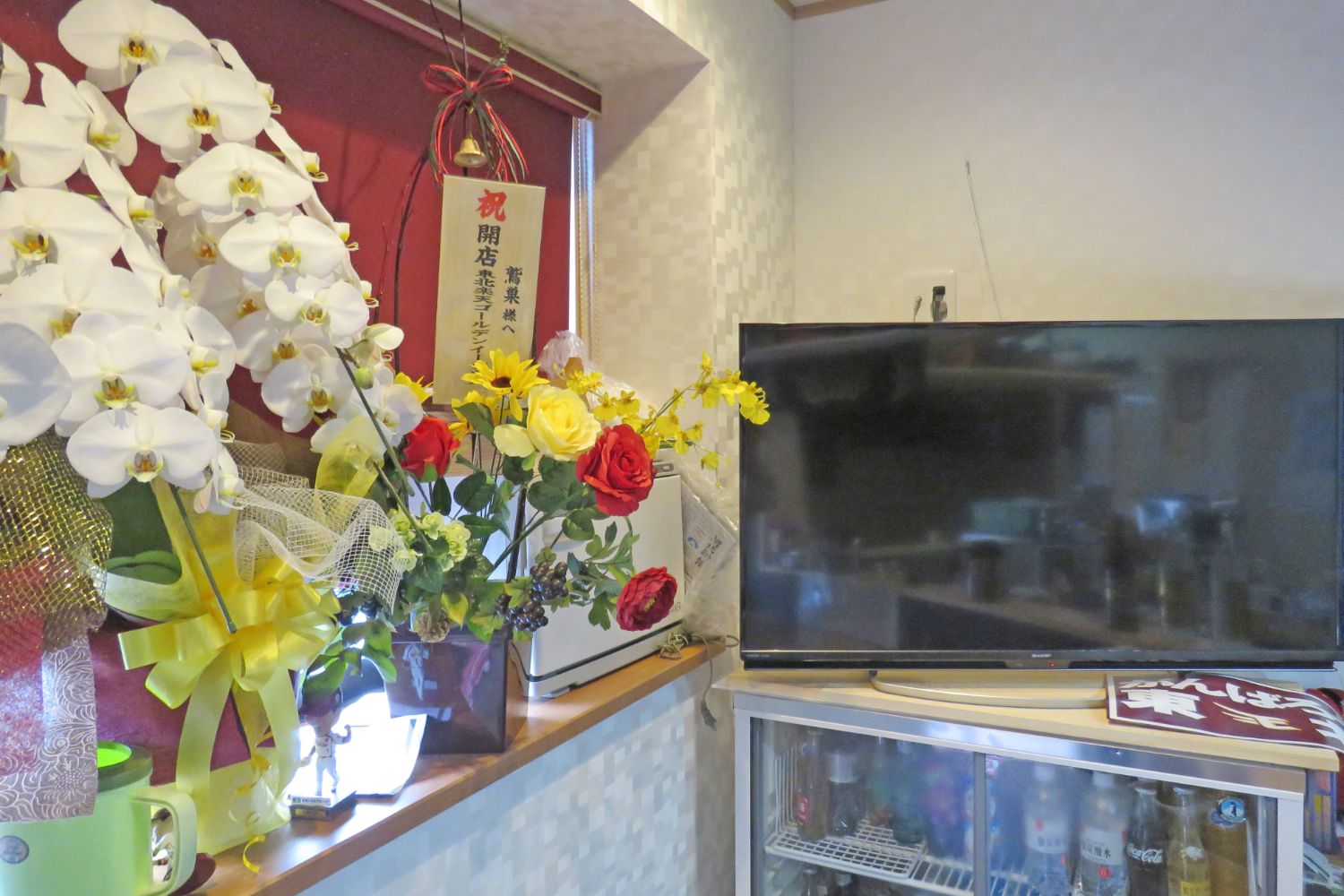 右側に飲み物が入った冷蔵庫の上にテレビが設置され、左側には白い胡蝶蘭や開店祝いの札が立てられている赤・黄色のバラを含む花束が並べられた居酒屋の開店祝いの室内の様子を写した写真