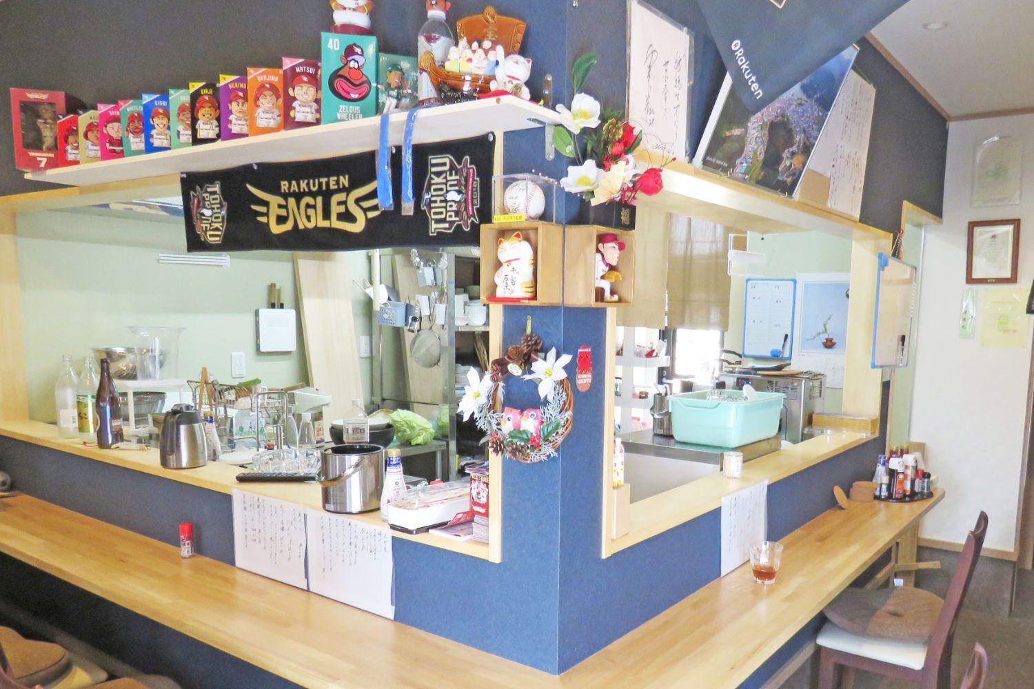 木製のL字型のカウンターとイスが並んだカウンター上には調味料類が置かれ、奥には調理器具や家電が見えるキッチンがあり、棚の上には「RAKUTEN EAGLES」のタオルや小さなフィギュアなどが飾られ、壁やカウンターには手書きのメモやメニューが貼られている居酒屋イーグルスの店内の写真