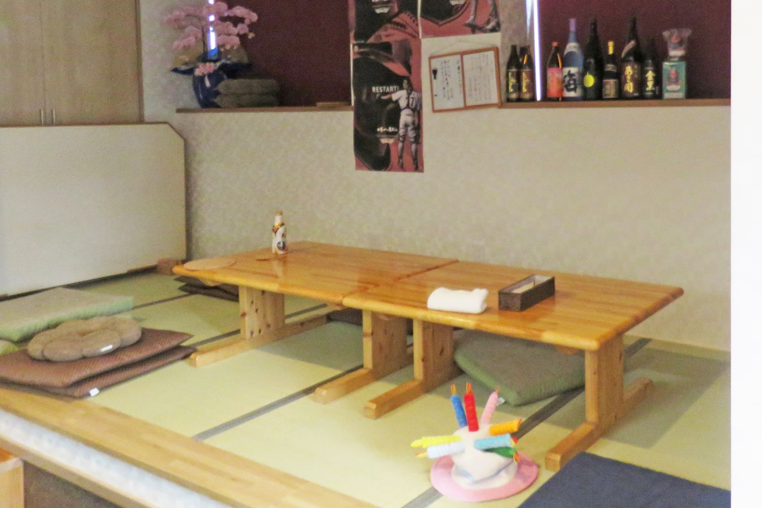 背景の壁にはポスターが貼られ、棚には複数のボトルが並べられている畳敷きの床に箸入れや布巾などが置かれた低い木製のテーブルと座布団が配置された店内を写している、居酒屋イーグルスの座敷席の写真