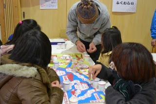 テーブルに広げたすごろくを囲んで子どもたちが紙コップをこまに使って遊びを楽しんでおり、スタッフがルールを説明している写真