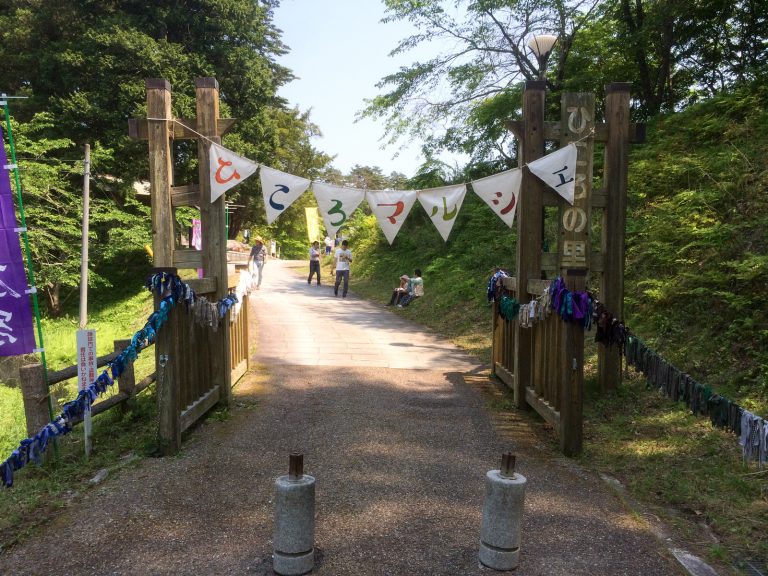 山間の小道の入り口にある木製の門に、ひらがなで「ひころマルシェ」と書かれたフラッグガーランドが掲げられ、人々がのんびりと歩いている様子の写真