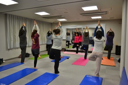 部屋の前面には大きな鏡が設置された室内でカラフルなヨガマットが敷かれ、片足を上げてもう一方の足の内ももに乗せ、両手を頭上で合わせた同じポーズをとっているストレッチの教室の様子を写した写真