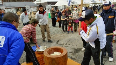 周囲で観客が見ている中、白色の上着を着た男性が杵を持ち、ニット帽を被った女性が手を添えてともちつきをしている様子の写真