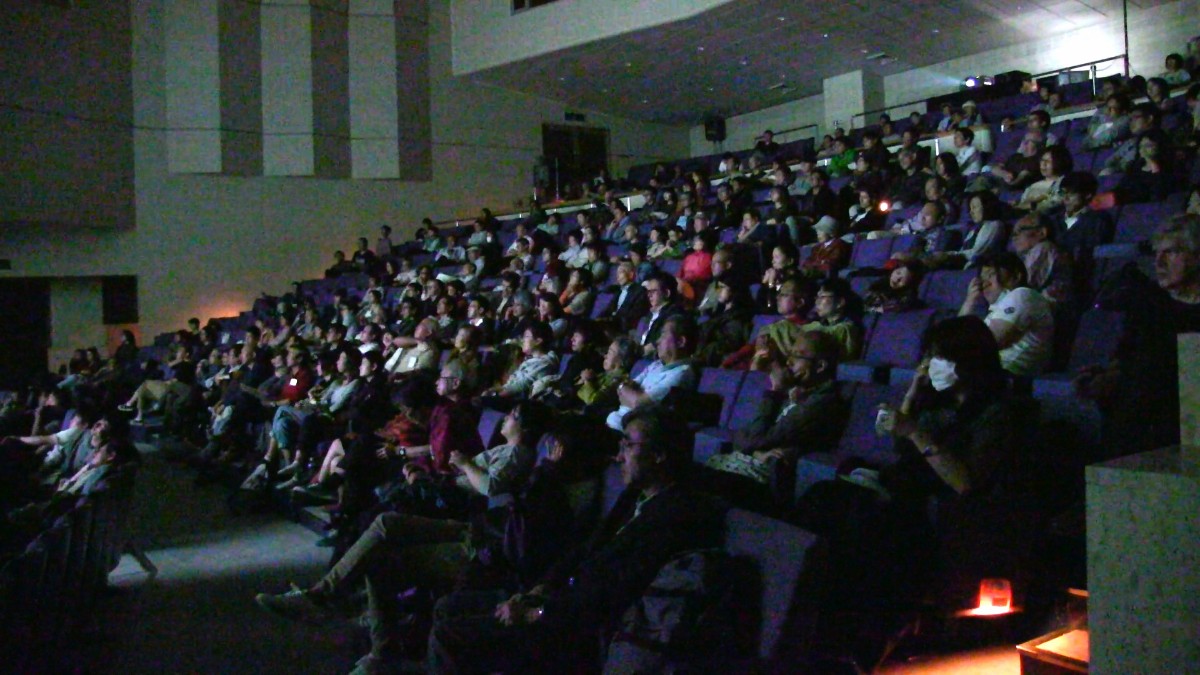 映画『願いと揺らぎ』の上映中に多くの観客が座って真剣にスクリーンを見つめている暗い劇場内の様子を写した写真。