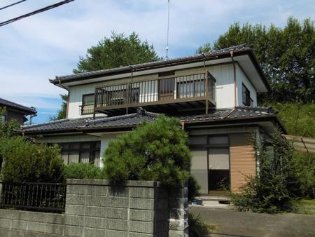 手前に塀と植木があり二階に長いベランダがある白い外壁と黒い瓦屋根を持つ二階建て住宅の写真