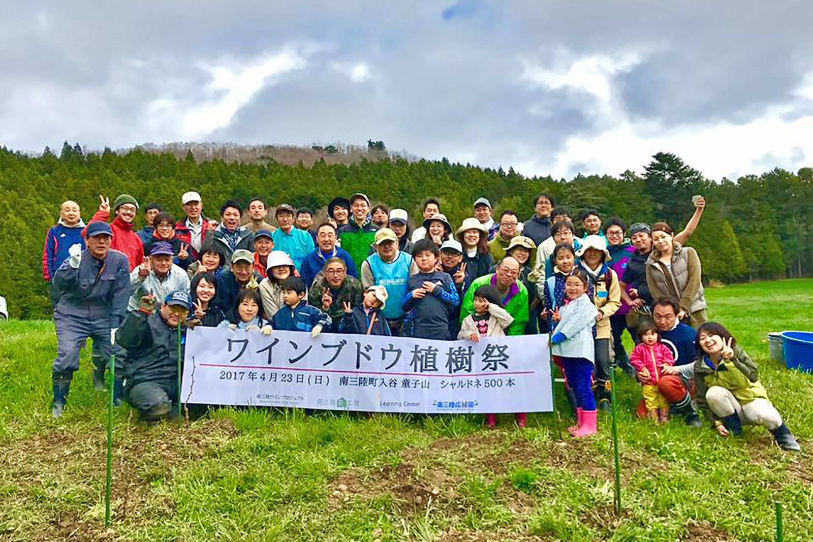 緑の森を背景に、草の斜面に並んだ大勢の参加者たちが「ワインブドウ植樹祭」と書かれた横断幕を掲げて写した集合写真