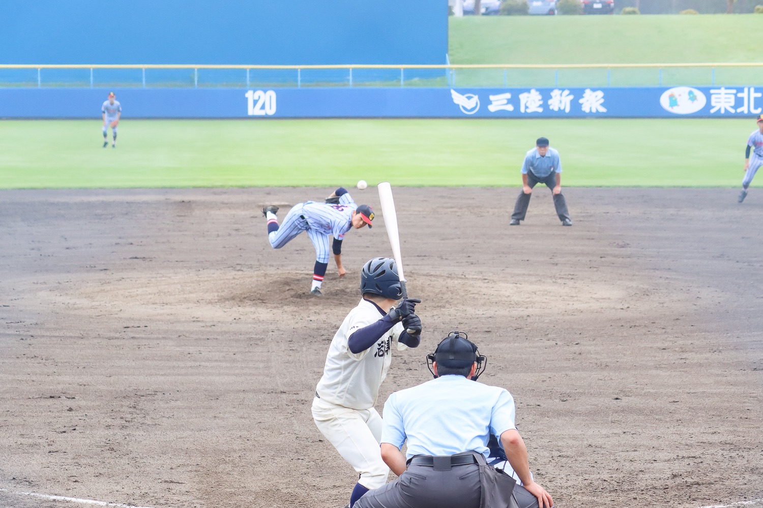キャッチャーと審判が構え、外野には守備が広がっているなか、縦縞のユニフォームを着たピッチャーが全力でボールを投げ、白いユニフォームのバッターが打とうとしている、緊張感のある瞬間を捉えた高校野球の投球シーンの写真