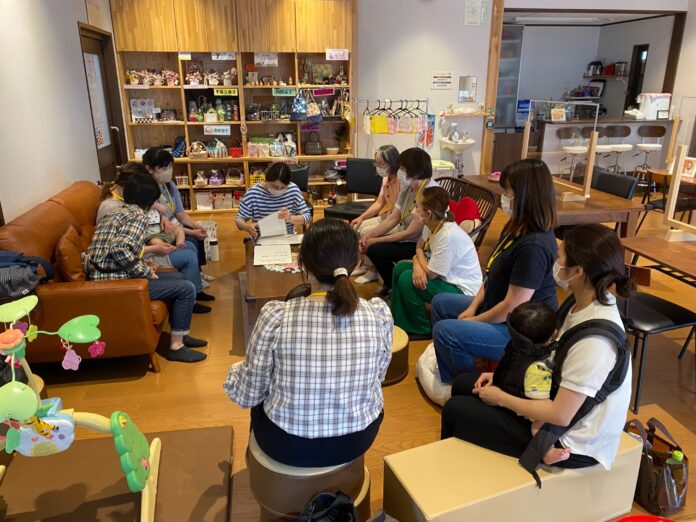 赤ちゃんを連れた母親たちがソファや椅子に座り、テーブルを囲んで「あずかりあいっこ」について真剣に話し合っている写真