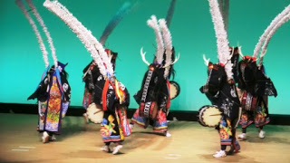 色鮮やかで華やかな衣装に身を包み、長い白い羽根飾りのついた大きな頭飾りをかぶった子どもたちが、太鼓を持って舞台の上で「行山流水戸辺鹿子躍(ぎょうざんりゅうみとべししおどり)」を披露している郷土芸能発表会の写真