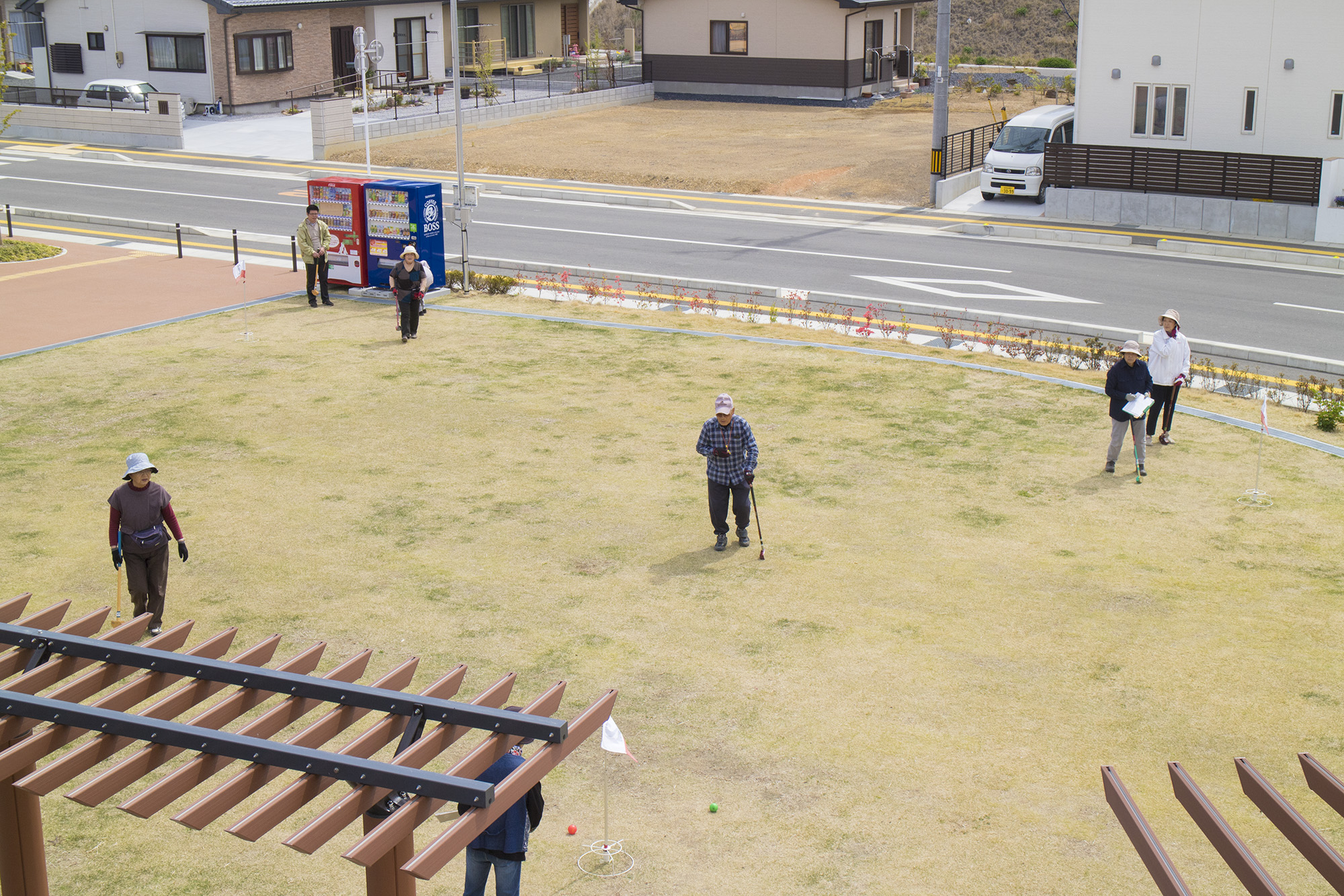 住宅街に面した芝生の広場で、複数の高齢者たちがグラウンドゴルフを楽しんでおり、スコアを記録する人やプレーに参加する人たちが点在している様子を高い位置から撮影した写真