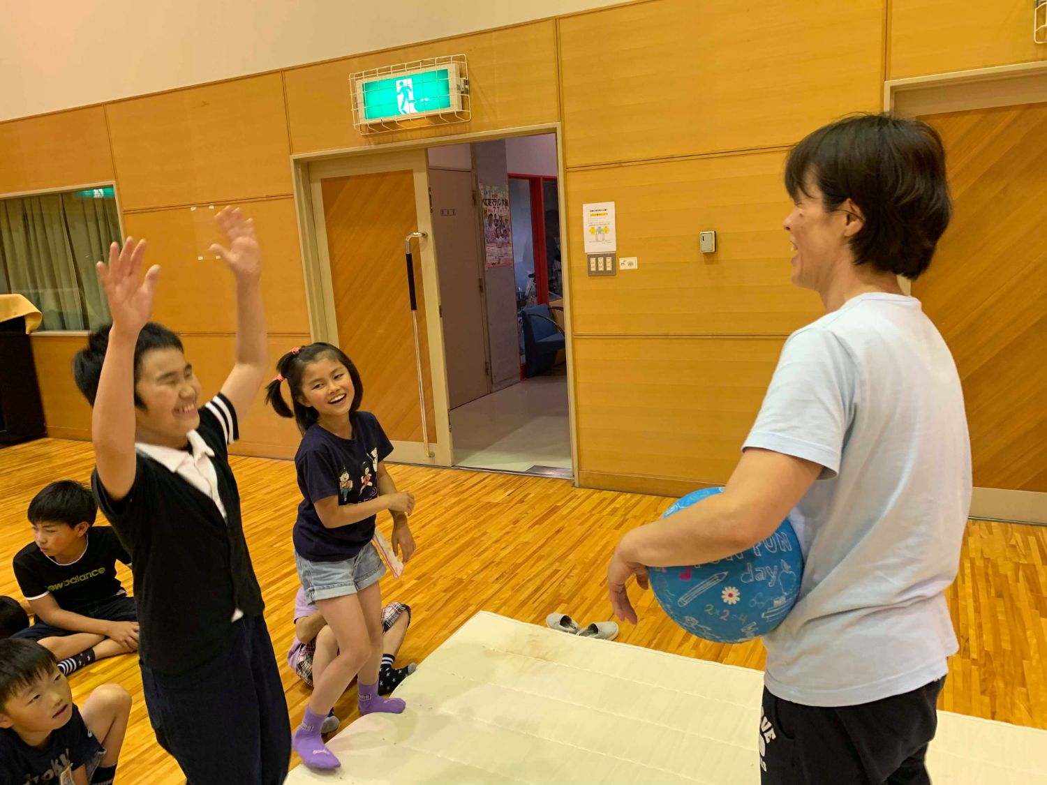 体育館の床に敷いたマットの傍に他の子供たちが座り、青いボールを持って立っているTシャツ姿の女性と向かい合ってマットの上に立つ笑顔の2人の子どもの内、男の子が両手を上げている写真