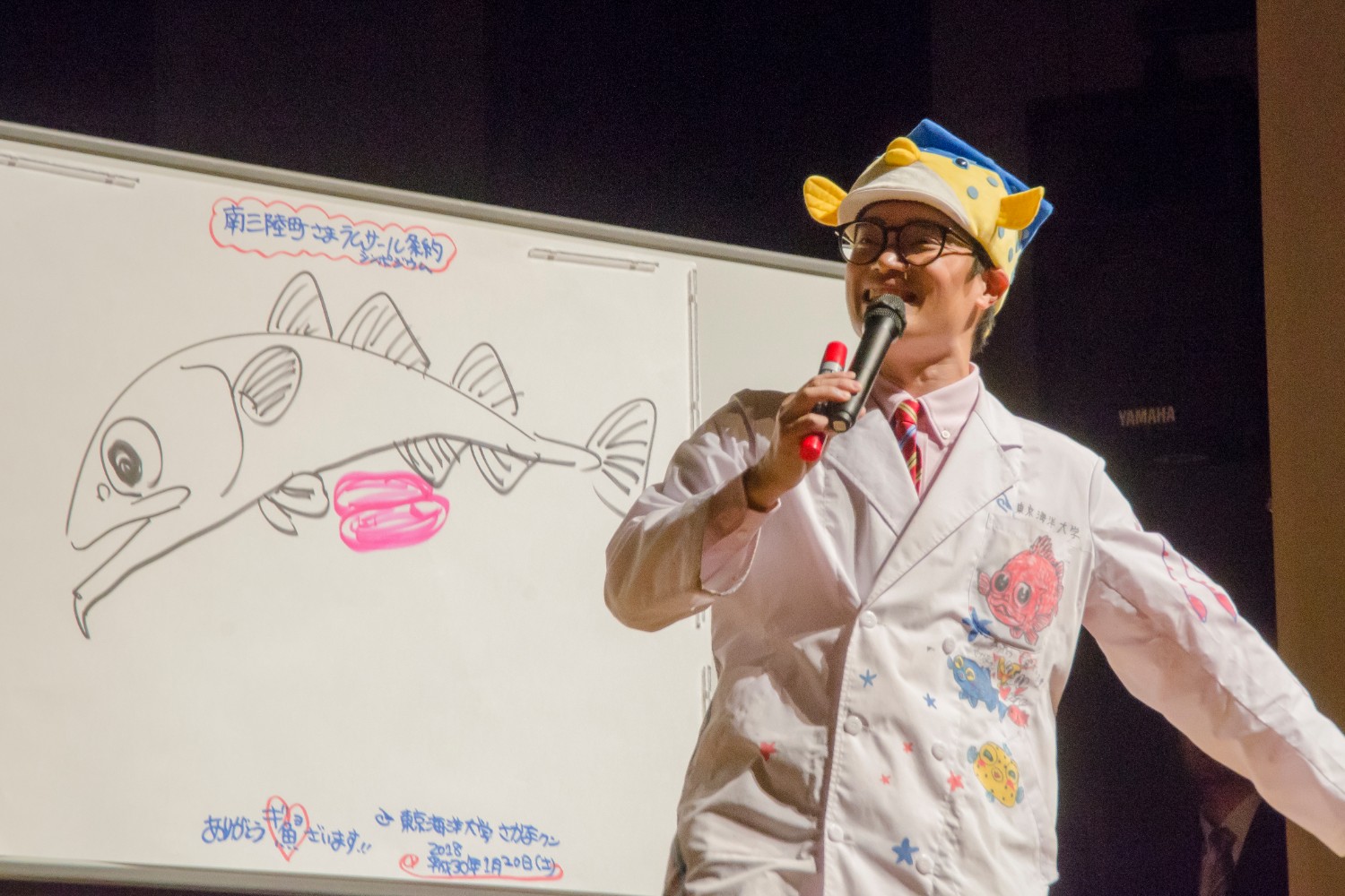 ホワイトボードに魚の絵を描きながら、マイクを持って話をしているさかなクンの写真