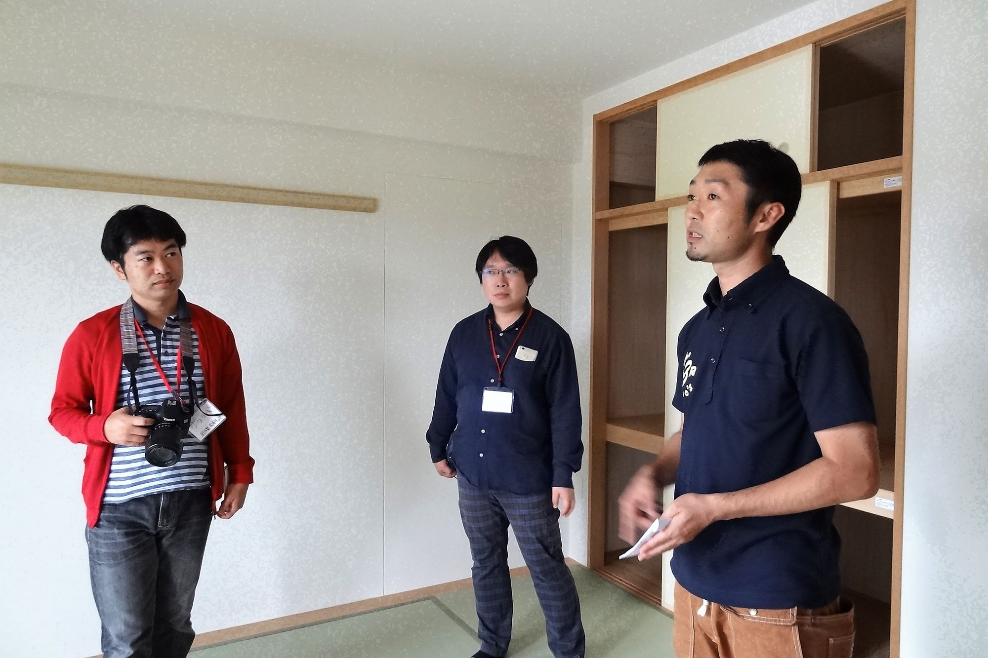 和室の一角で3人の男性が立ち話をしており、左の男性はカメラを首から下げ、中央の男性は手を下げて正面を見つめ、右の男性が説明している様子の写真