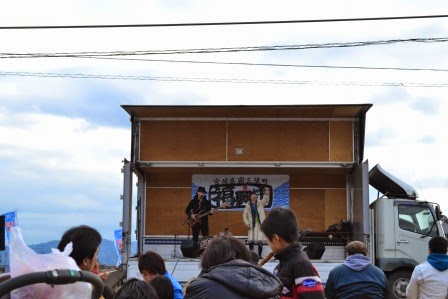 トラックの荷台が即席のステージになっていて、音楽演奏が行われている様子を観客が見守っている写真