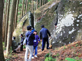 白い苔や模様がついた大きな岩が森の中に立っていて、その岩の前で数人の子どもと大人が観察したり触れたりしながら過ごしている様子の写真