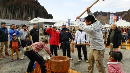 人々が見守るなか、白い上着の男性が高く振り上げた杵で臼の中のもち米をつこうとしており、隣でニット帽をかぶった女性がタイミングを見て手を添えようとしている様子を、背景に屋台やのぼり旗が並ぶ冬の野外会場で撮影した写真