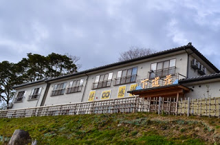 斜面の上に建つクリーム色の外壁の二階建ての建物が、木々の背景とともに写されている民宿の全景の写真