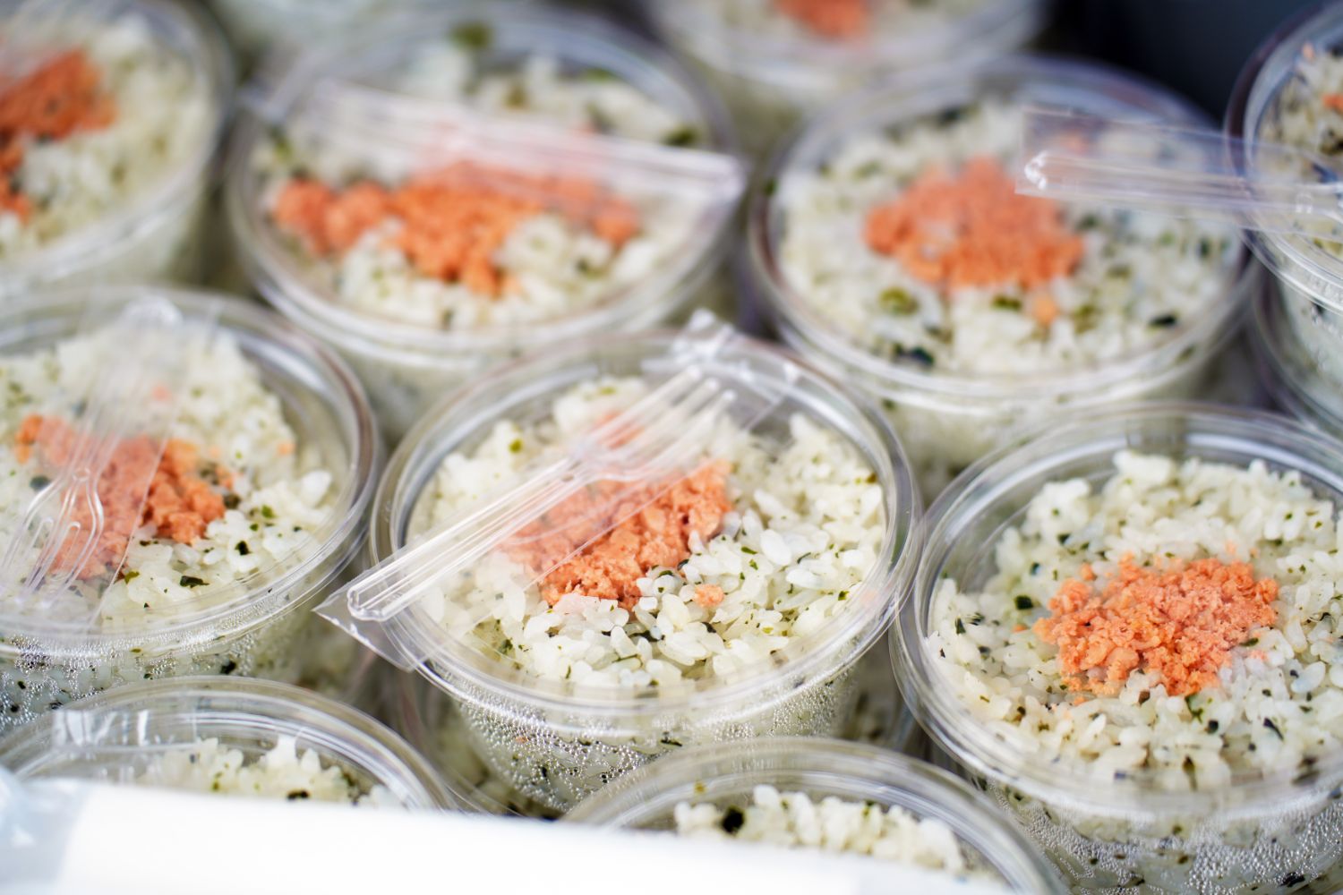 透明なカップに盛り付けられた鮭のフレークとわかめが入ったごはんが整然と並べられ、上には使い捨てフォークが添えられている、ツール・ド・東北の補給食として提供された鮭わかめごはんの写真