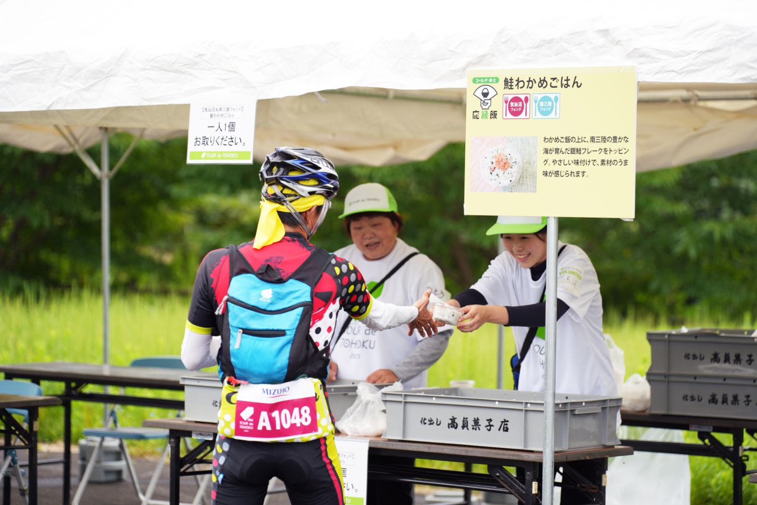 サイクルジャージを着た参加者が、エイドステーションのテントで笑顔のボランティアスタッフから鮭わかめごはんを受け取っている、ツール・ド・東北の補給所での給食提供の様子の写真