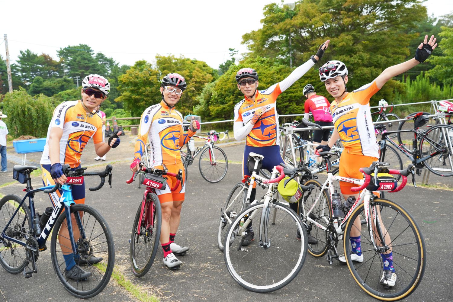 オレンジと白のチームジャージを着た4人の参加者が、自転車のそばで笑顔を見せながら親指を立てたりポーズをとったりしている、ツール・ド・東北のスタートでの集合写真