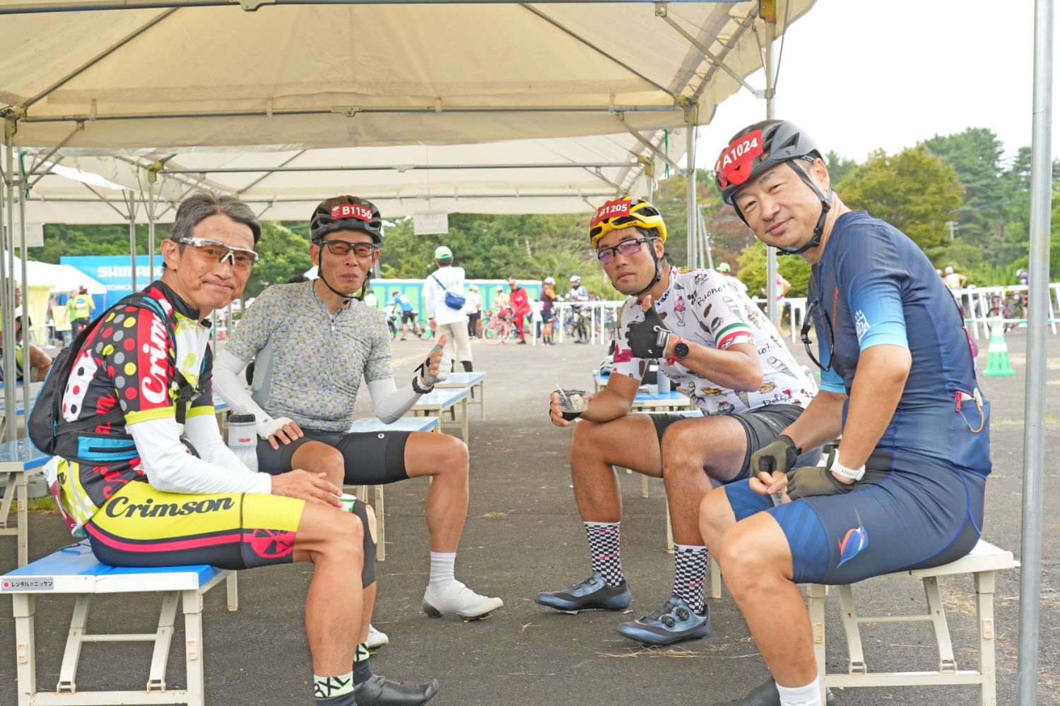 4人の男性サイクリストが自転車ウェアとヘルメットを着用し、ベンチに座って休憩しながらカメラに向かって親指を立てたり微笑んだりしている様子の写真