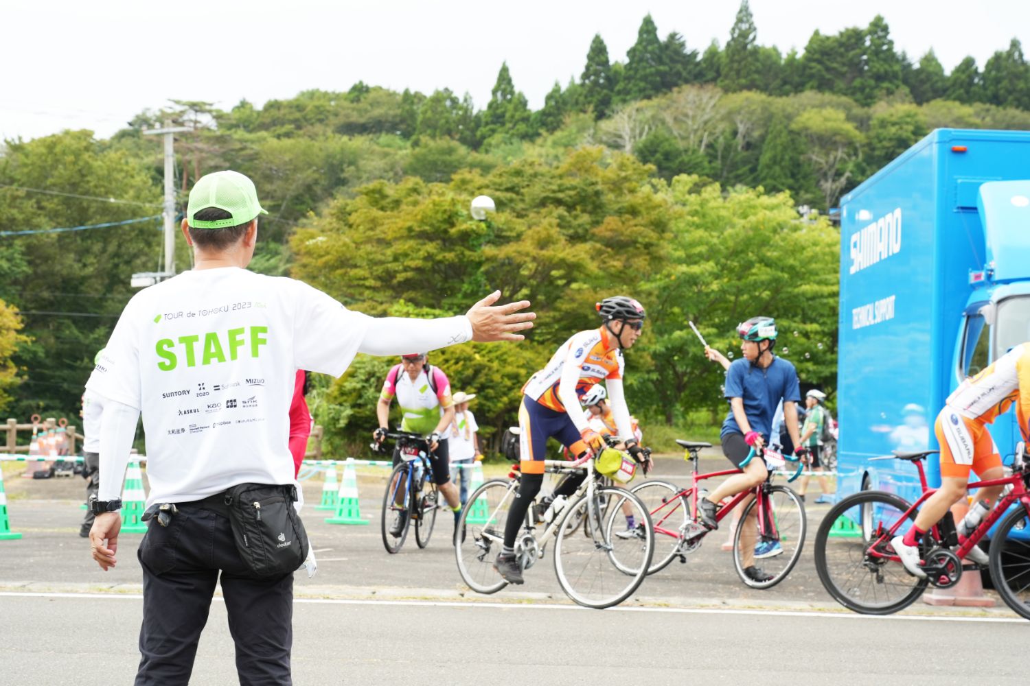 白いスタッフTシャツを着た誘導員が腕を広げて参加者を案内し、複数のサイクリストが自転車にまたがって動き出している、ツール・ド・東北の運営と参加風景の写真