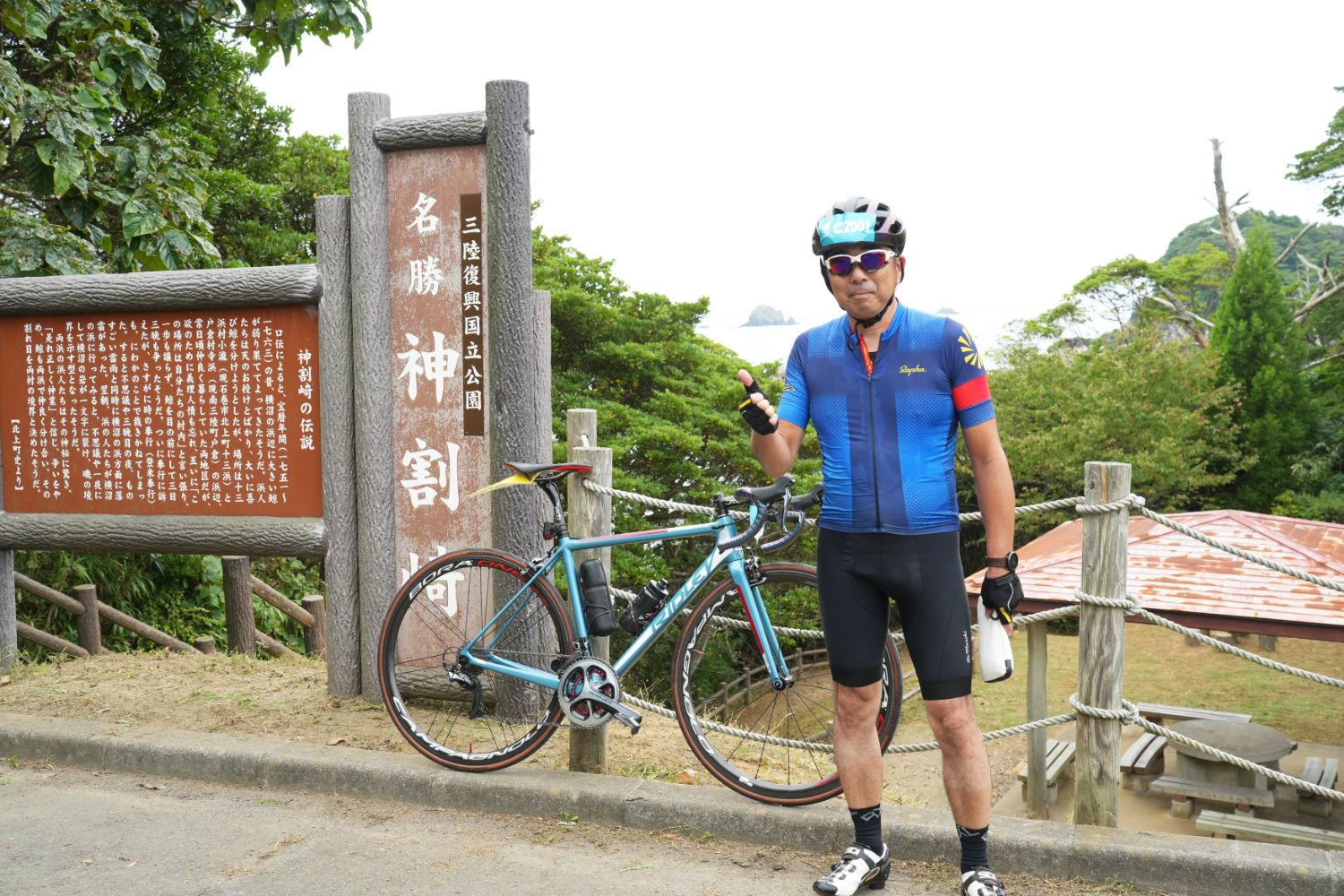 三陸復興国立公園の神割崎の石碑前で、青いサイクルジャージを着た男性サイクリストが自転車を立てかけてカメラに向かって親指を立てている様子の写真