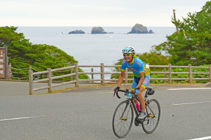 海に浮かぶ岩と木製の柵を背景に、青と緑のサイクルジャージを着た参加者が笑顔でロードバイクをこぎながらツール・ド・東北のコースを走行している様子の写真