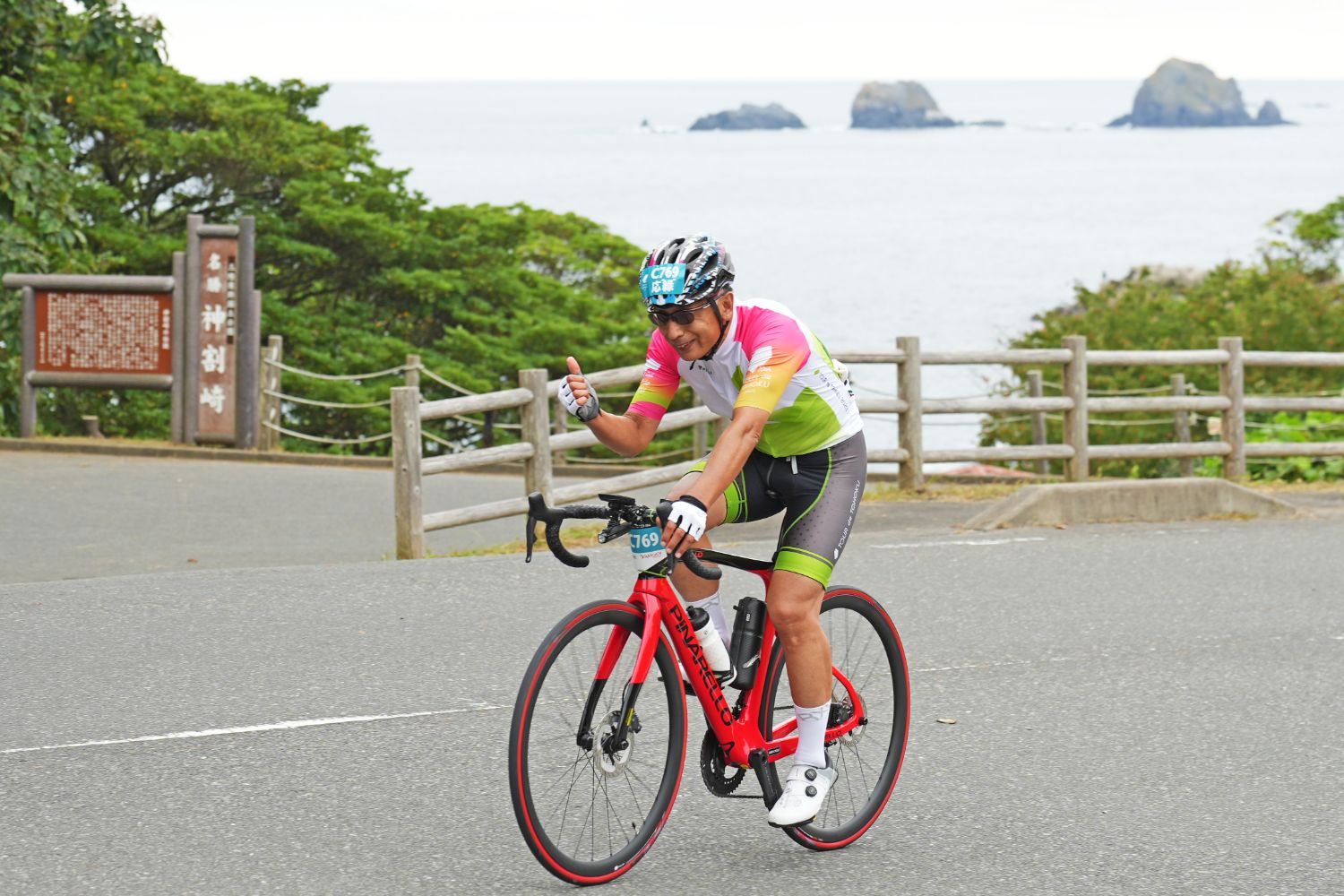海を背景にした道路を赤いロードバイクで走行中の男性サイクリストが、片手で親指を立てて笑顔を見せている様子の写真