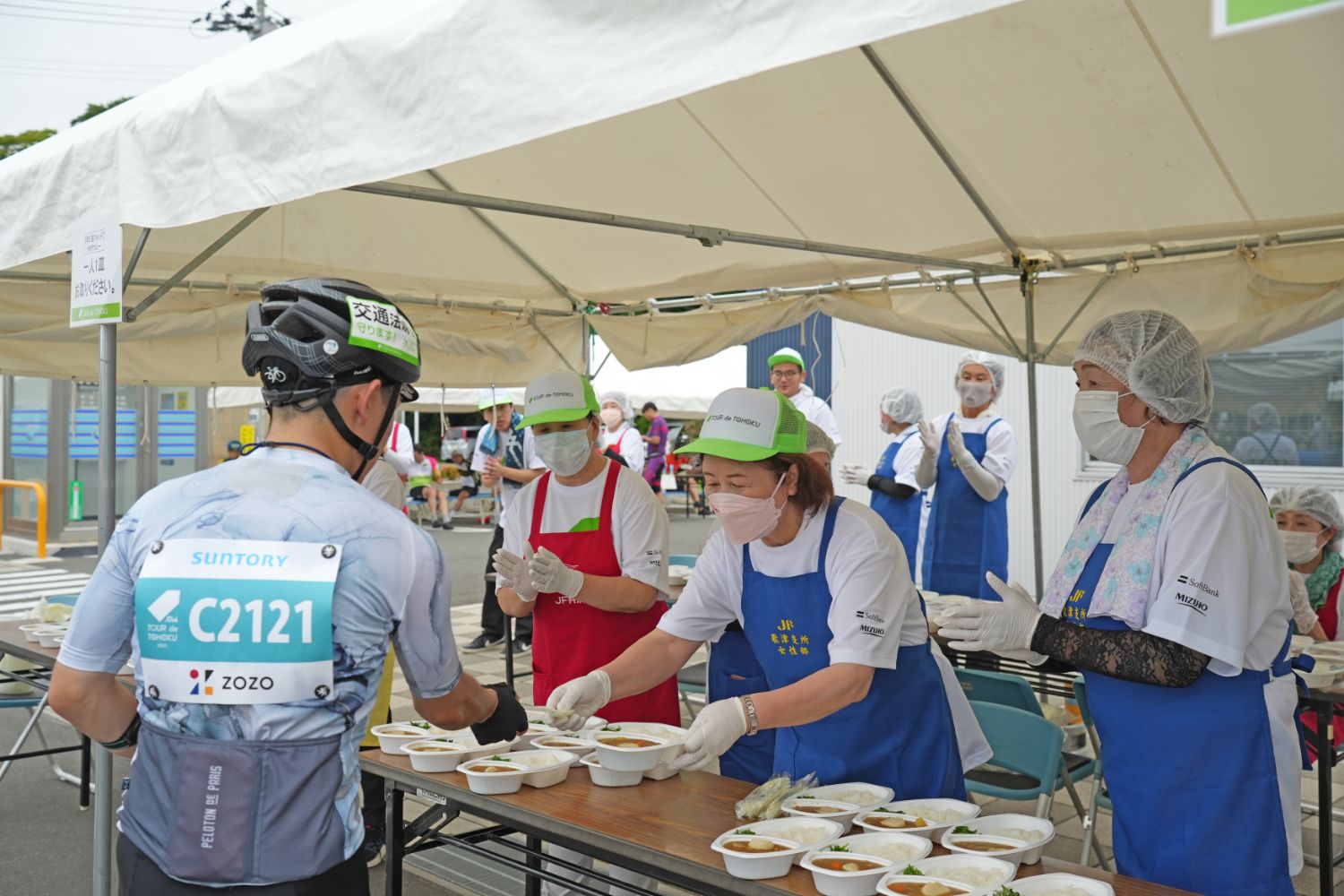 自転車レースに参加している選手が、給食テントでスタッフから食事を受け取っている様子の写真