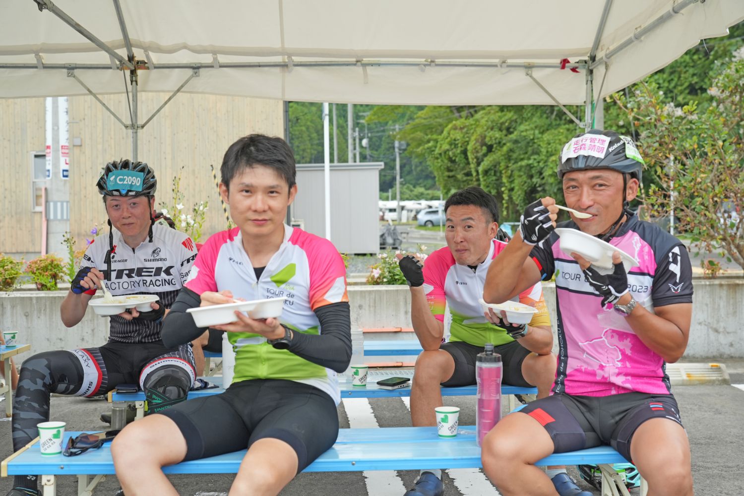 休憩所のテントの下で、サイクリング中の男性4人がユニフォーム姿のままベンチに座り、笑顔で食事を楽しんでいる様子の写真と