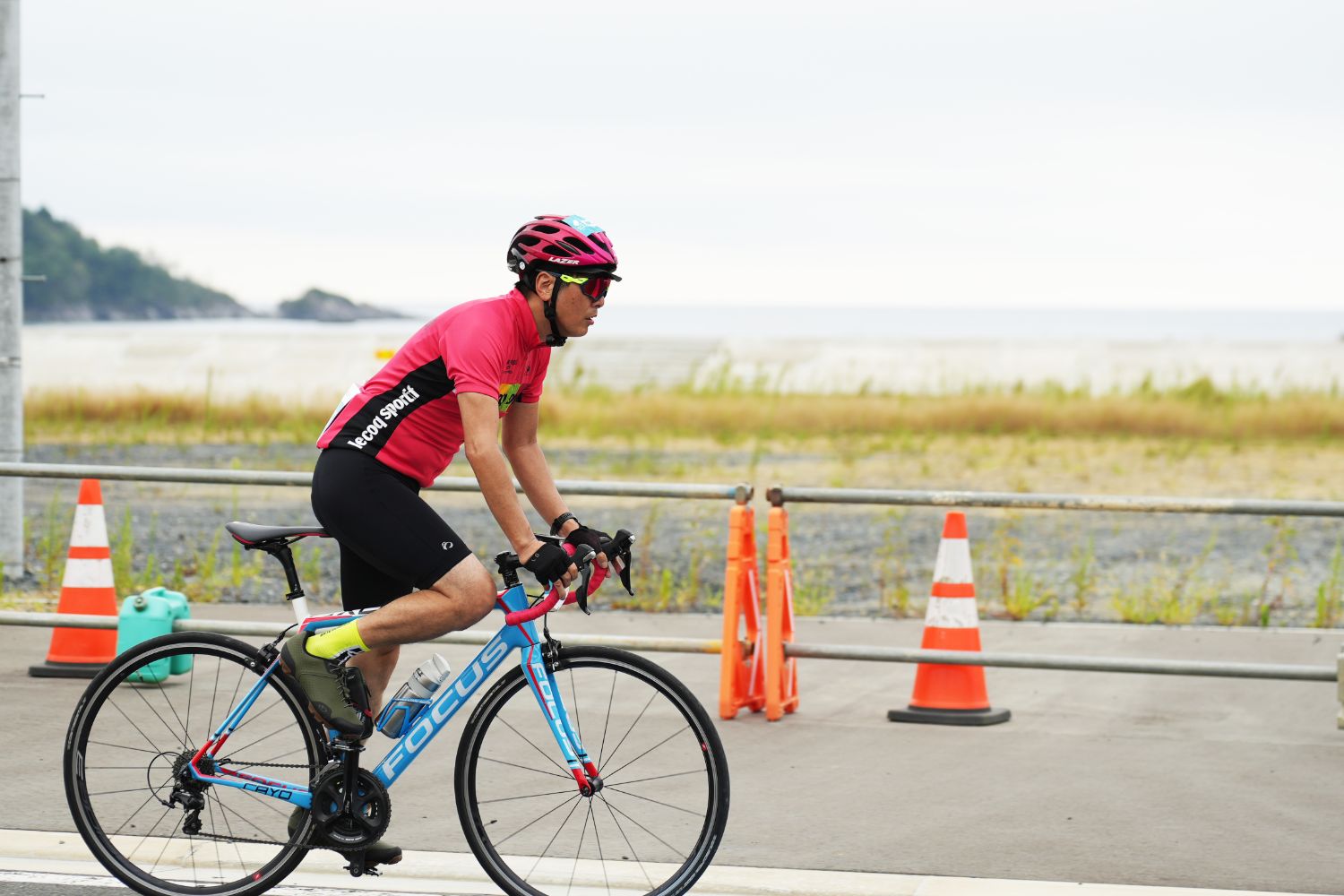 海沿いの道路を、ピンク色のユニフォームを着たサイクリストが一人で自転車に乗って走行している様子の写真