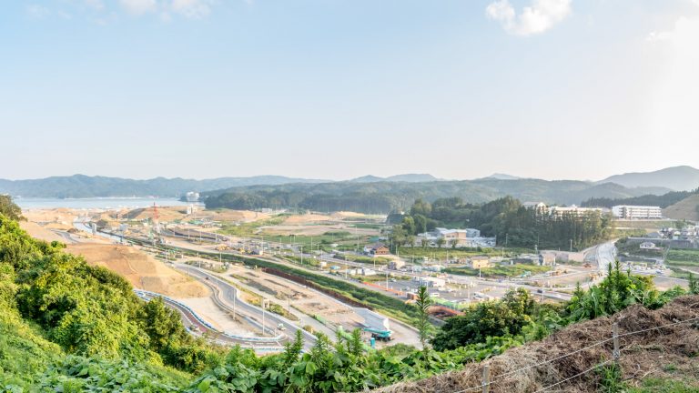 山並みや海を背景に、川沿いの右側には建物が立ち並び、左側には工事車両があり造成が進む町の様子を高台から撮影した写真