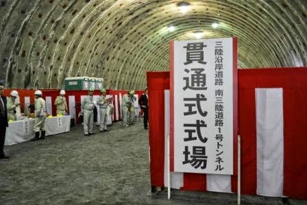 「貫通式式場 三陸沿岸道路 南三陸道路1号トンネル」と書かれた大きな看板が掲げられた、紅白幕が張られた曲線天井の地下トンネル内に、白いヘルメットと作業服を着用した人々が集まっている写真