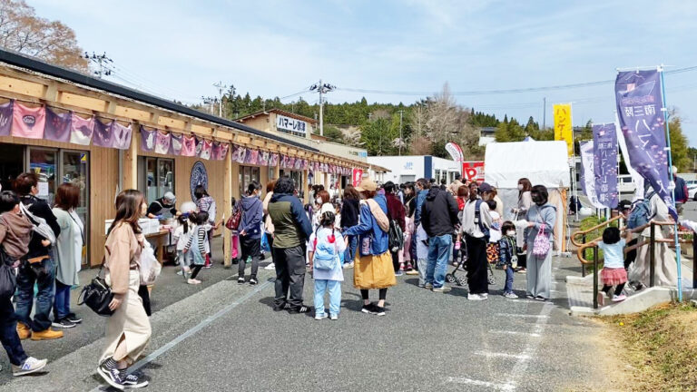 紫色ののぼり旗が並ぶ木造の建物の前で、子どもから大人まで多くの人々が屋台やブースに集まり、行列を作ったり交流したりするにぎやかな祭りの様子を写した写真