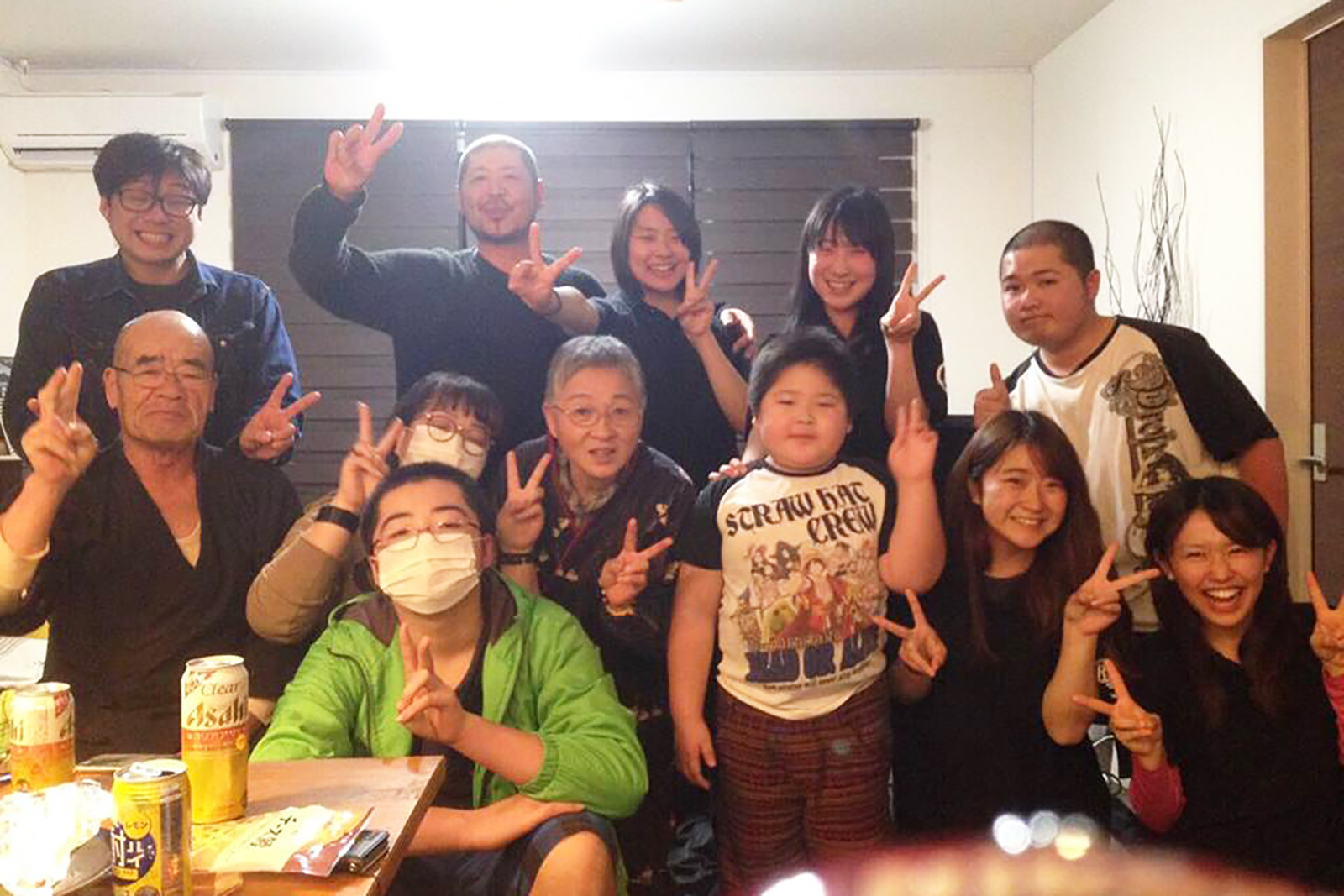 室内で若い人から年配の方、子どもまで幅広い世代の男女が笑顔で集まり、ピースサインをしている集合写真